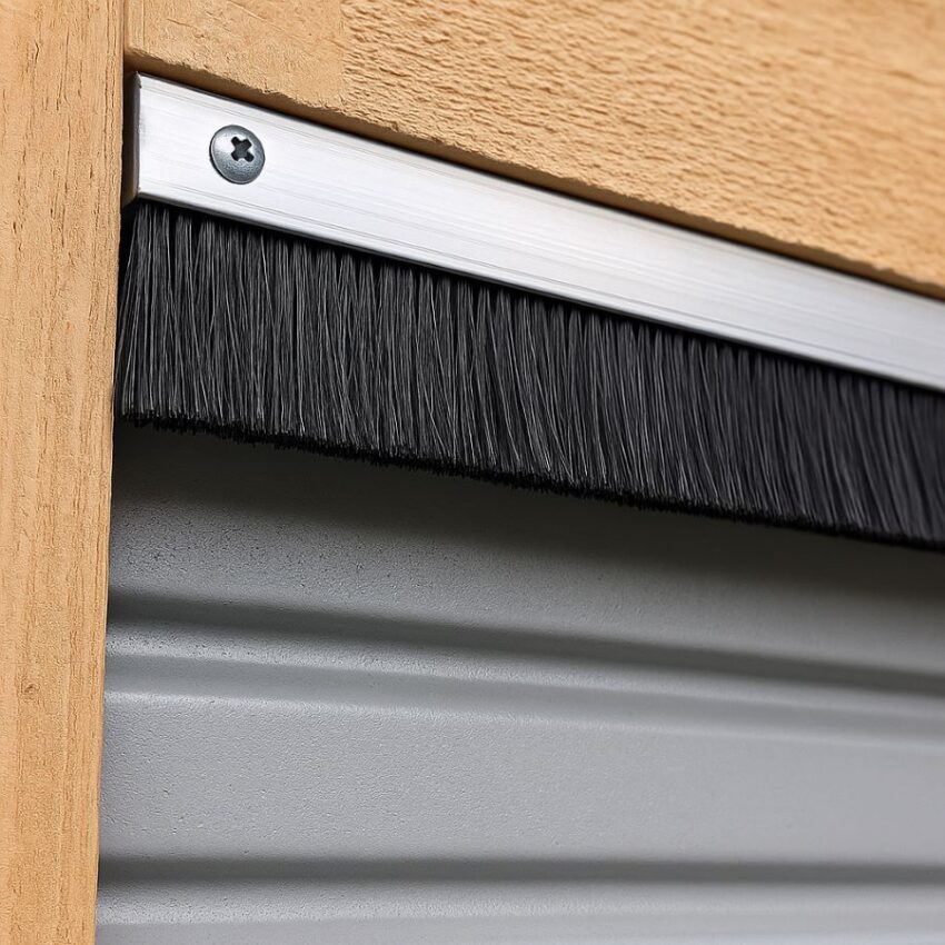 Close‑up view of a brush seal with straight aluminum holder mounted along the top of a roll‑up garage door frame, bristles touching the door edge