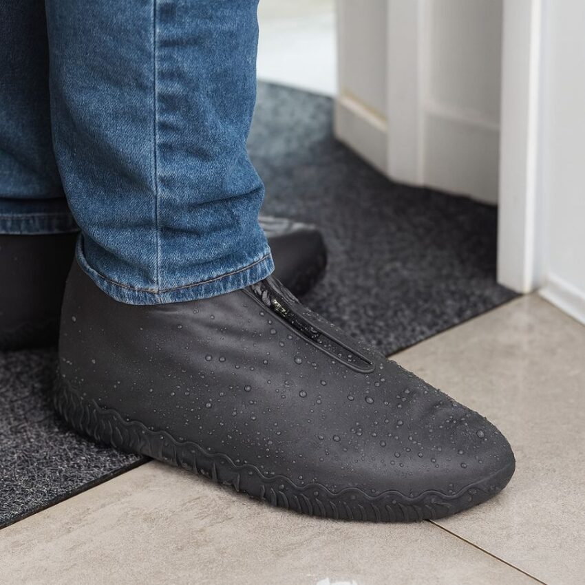 Person standing on an entry mat wearing gray zippered waterproof shoe covers with water beading on the surface.